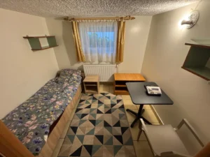 Pokój jednoosobowy z łóżkiem, stolikiem, krzesłem i półkami na ścianie
